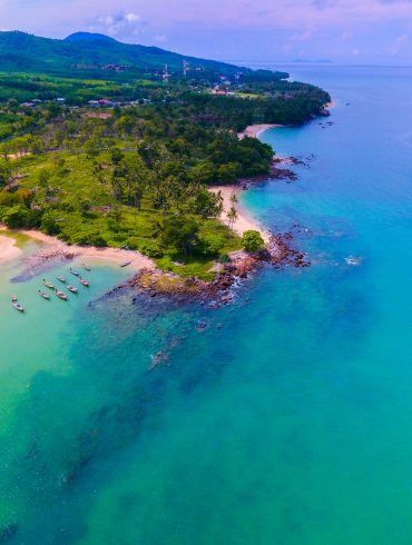 Love London: A scenic aerial view of Koh Samui's tropical coastline showcases turquoise waters meeting a sandy beach. Lush greenery and hills in the background paint a stunning picture, with a few boats anchored near Bo Phut's shore under a clear sky dotted with scattered clouds.