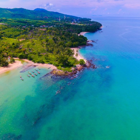 Love London: A scenic aerial view of Koh Samui's tropical coastline showcases turquoise waters meeting a sandy beach. Lush greenery and hills in the background paint a stunning picture, with a few boats anchored near Bo Phut's shore under a clear sky dotted with scattered clouds.