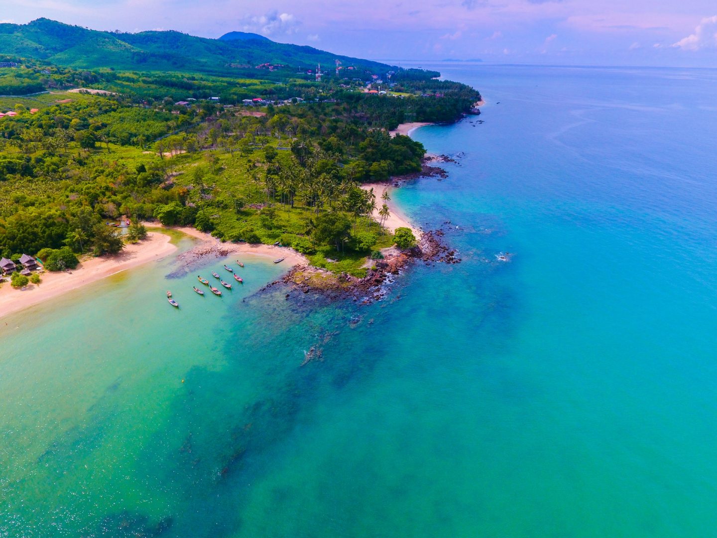 Love London: A scenic aerial view of Koh Samui's tropical coastline showcases turquoise waters meeting a sandy beach. Lush greenery and hills in the background paint a stunning picture, with a few boats anchored near Bo Phut's shore under a clear sky dotted with scattered clouds.