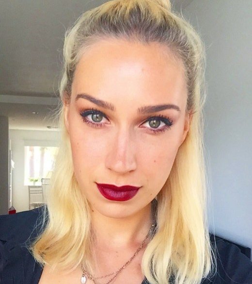 Love London: A person with long blonde hair in a top knot poses indoors, showcasing a Swedish look. They are wearing bold red lipstick and dark eye makeup, exuding elegance with their neutral expression. The softly lit room in the background sets the perfect stage for a chic makeup tutorial.