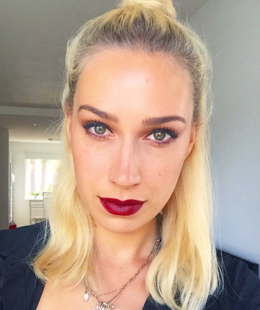 Love London: A person with long blonde hair in a top knot poses indoors, showcasing a Swedish look. They are wearing bold red lipstick and dark eye makeup, exuding elegance with their neutral expression. The softly lit room in the background sets the perfect stage for a chic makeup tutorial.