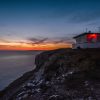 Love London: At sunset, the Dorset cliffside is magical—a small building glows red at its edge. The deep blues and orange hues of the sky reflect on the calm sea below, creating an idyllic staycation setting.