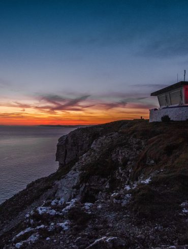 Love London: At sunset, the Dorset cliffside is magical—a small building glows red at its edge. The deep blues and orange hues of the sky reflect on the calm sea below, creating an idyllic staycation setting.