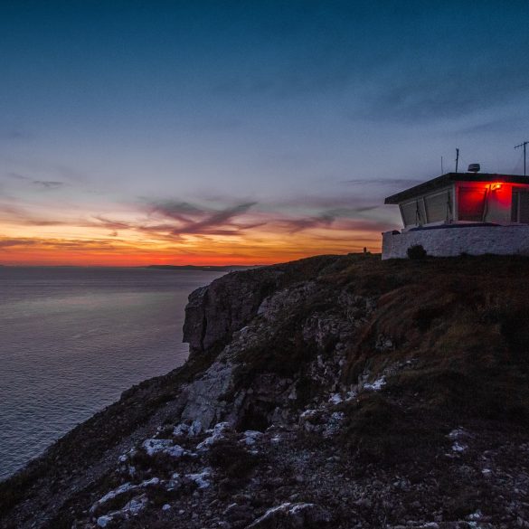 Love London: At sunset, the Dorset cliffside is magical—a small building glows red at its edge. The deep blues and orange hues of the sky reflect on the calm sea below, creating an idyllic staycation setting.
