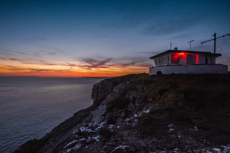 Love London: At sunset, the Dorset cliffside is magical—a small building glows red at its edge. The deep blues and orange hues of the sky reflect on the calm sea below, creating an idyllic staycation setting.