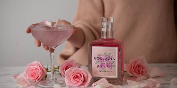 Love London: A person in a beige sweater holds a glass of pink gin cocktail garnished with petals, next to a bottle of pink collagen rose drink promising to help you look younger. Pink roses and petals are artfully scattered on the marble surface.