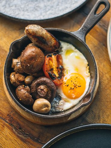 Love London: Enjoy a vibrant Electro Brunch with a breakfast skillet featuring a sunny-side-up egg, sausages, grilled tomato, and mushrooms. At Gaucho Broadgate, indulge in this delectable dish as part of their Bottomless Brunch spread on a rustic wooden table.