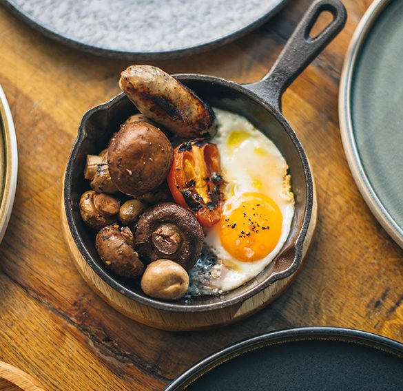 Love London: Enjoy a vibrant Electro Brunch with a breakfast skillet featuring a sunny-side-up egg, sausages, grilled tomato, and mushrooms. At Gaucho Broadgate, indulge in this delectable dish as part of their Bottomless Brunch spread on a rustic wooden table.