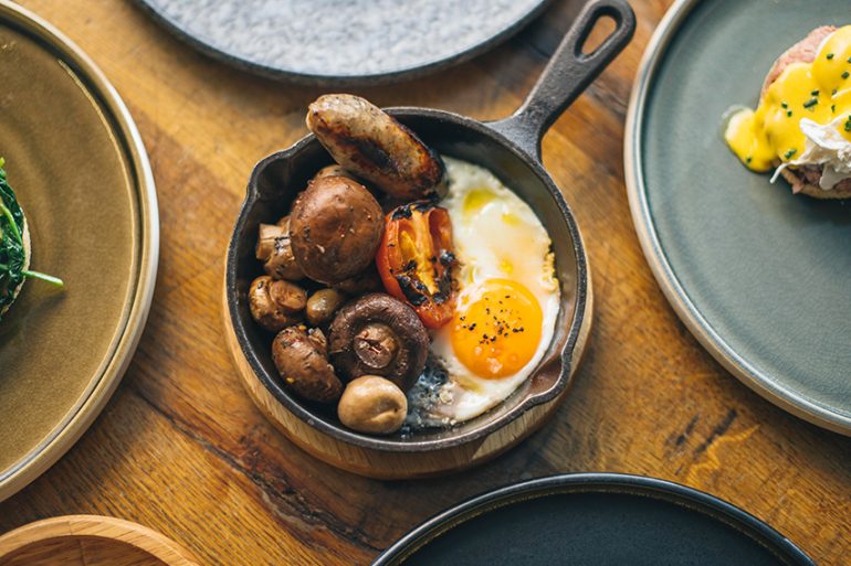 Love London: Enjoy a vibrant Electro Brunch with a breakfast skillet featuring a sunny-side-up egg, sausages, grilled tomato, and mushrooms. At Gaucho Broadgate, indulge in this delectable dish as part of their Bottomless Brunch spread on a rustic wooden table.