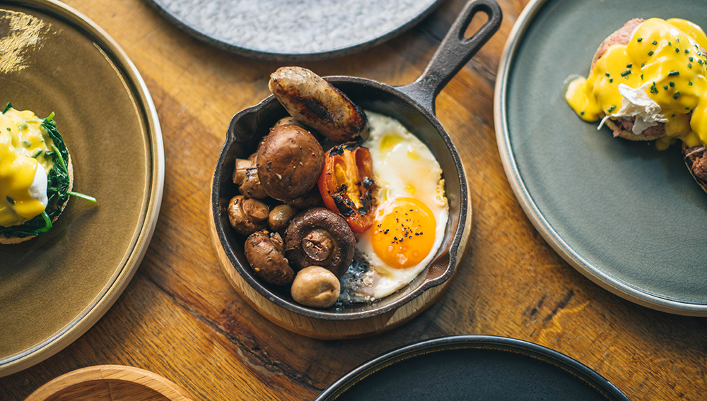 Love London: Enjoy a vibrant Electro Brunch with a breakfast skillet featuring a sunny-side-up egg, sausages, grilled tomato, and mushrooms. At Gaucho Broadgate, indulge in this delectable dish as part of their Bottomless Brunch spread on a rustic wooden table.