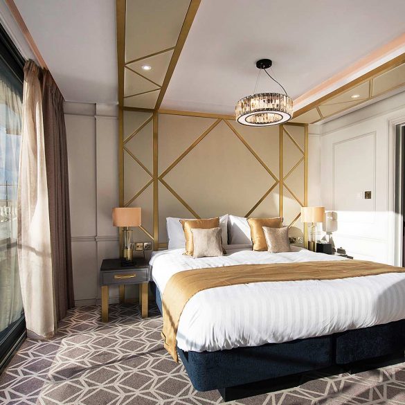 Love London: A luxurious Southend hotel room features a large bed, elegant gold and white decor, and a geometric patterned carpet. A large window showcases a scenic ocean view—a perfect reason to visit. A modern hanging light fixture and adjacent bathroom add to the opulent atmosphere.